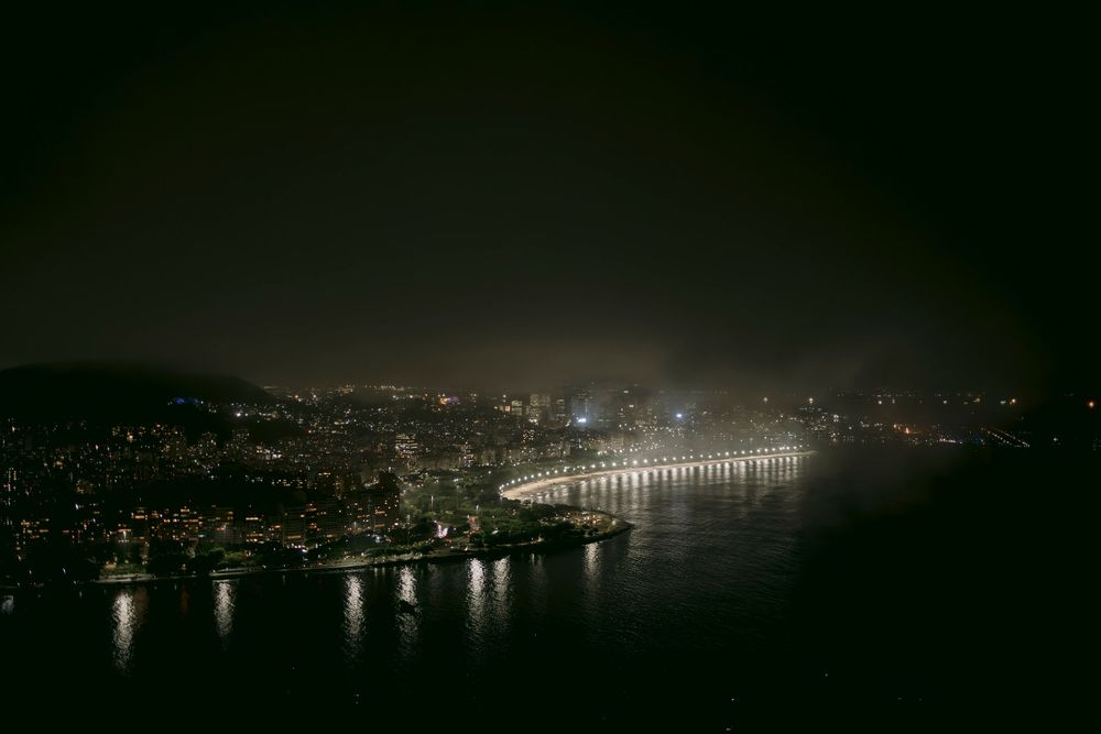Rio de Janeiro night skyline with curved coastal boulevard lights reflecting on dark bay waters.