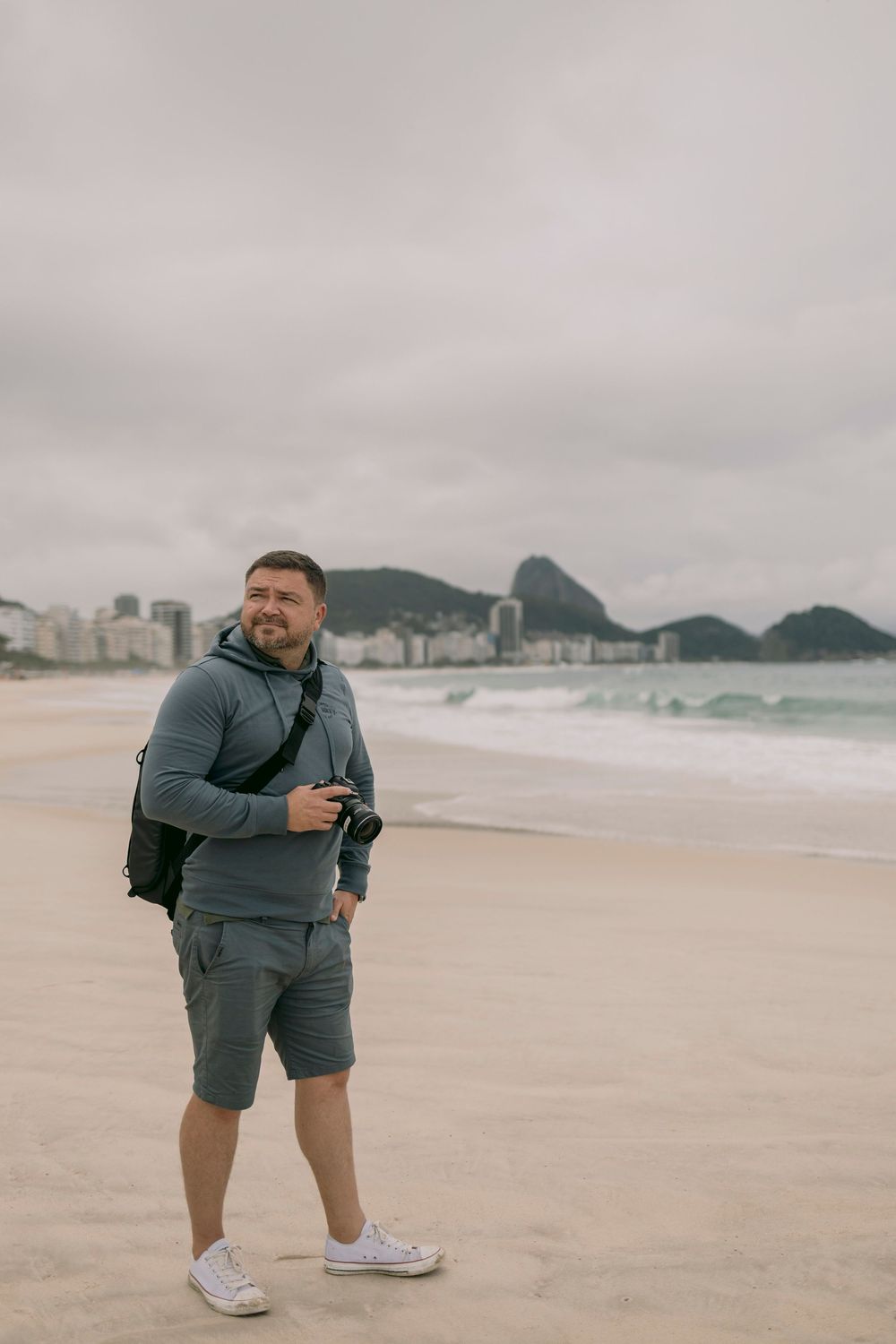 Smiling man holding camera on Copacabana beach with mountain backdrop in Rio de Janeiro.