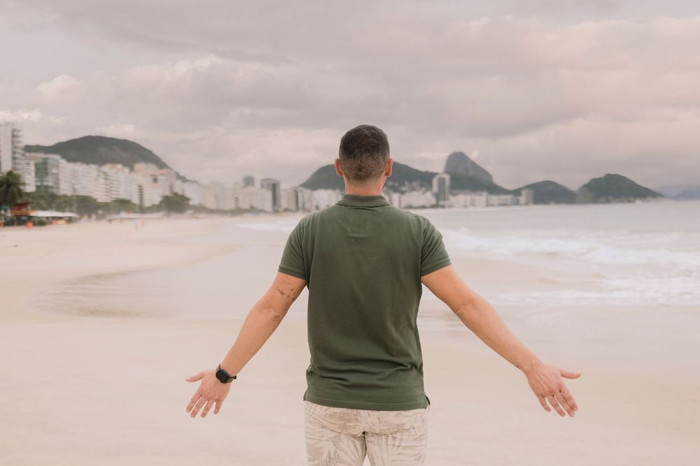 Man with outstretched arms facing Copacabana beach with Sugarloaf Mountain in background.