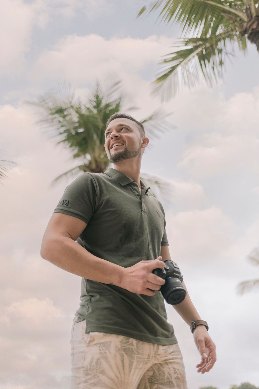 Smiling man holding a camera outdoors with palm trees and cloudy sky in background.