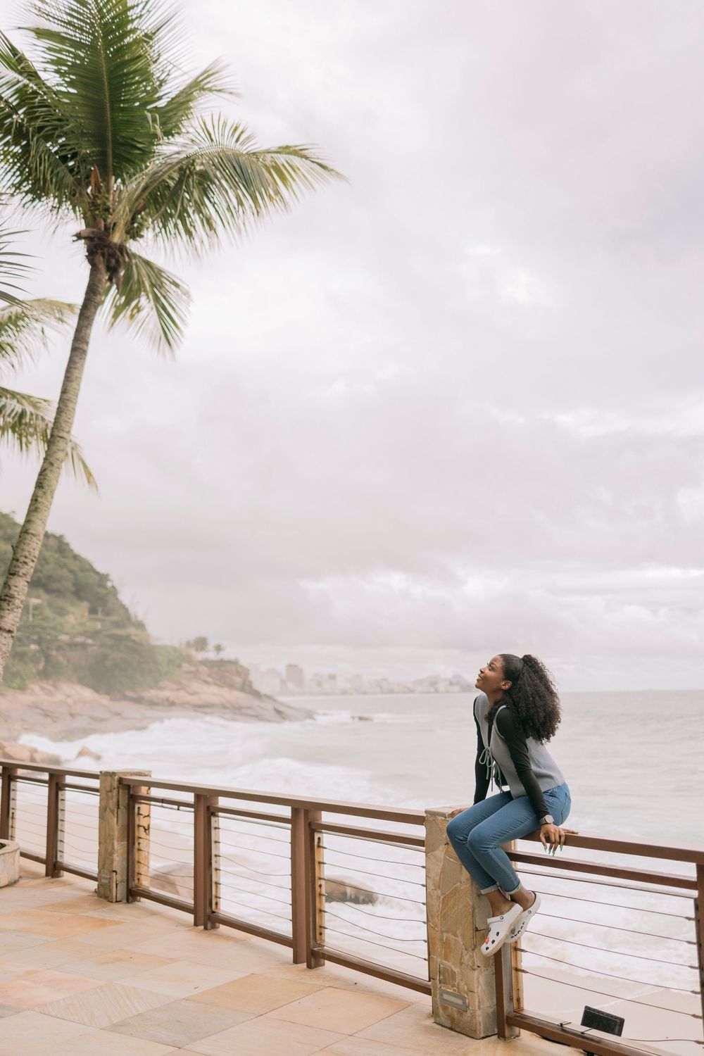 Woman sitting on railing by the ocean with palm tree and cloudy sky backdrop.