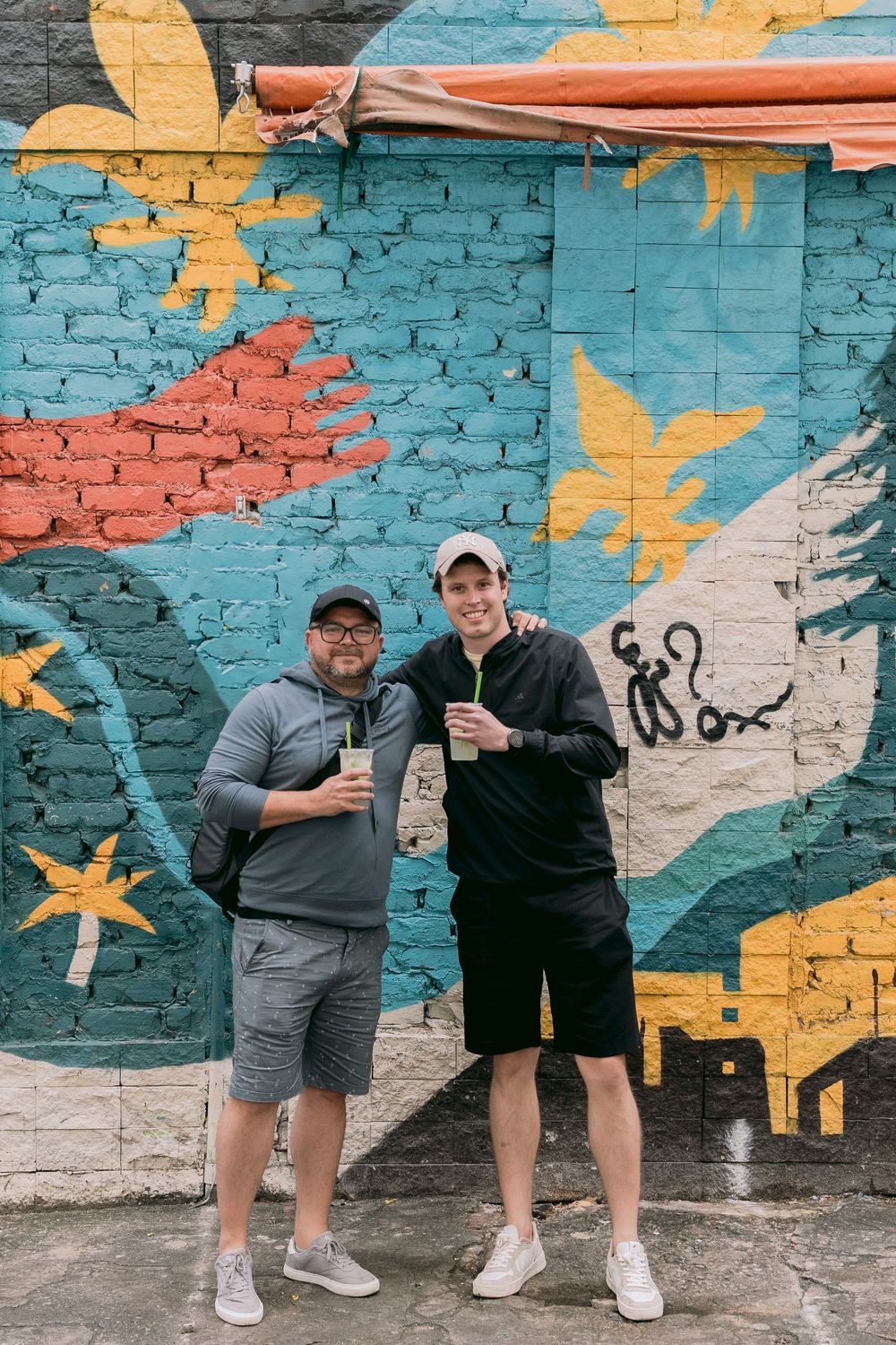 Two smiling men holding green drinks standing before colorful tropical street art brick mural.