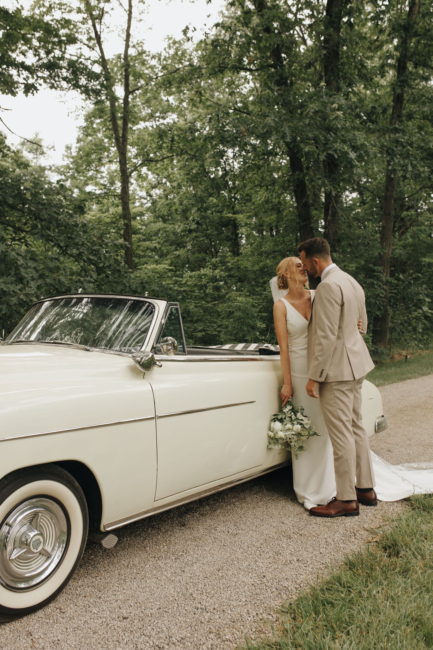 Bride and groom posing beside vintage car in front of tree-lined driveway at Athens, Ohio backyard wedding