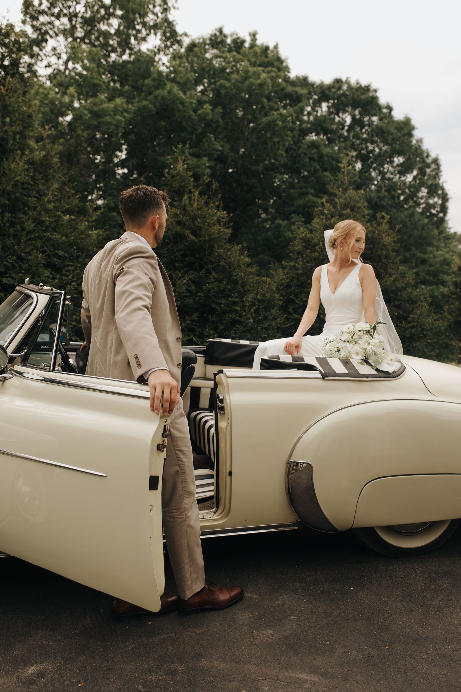 Editorial-style photo of bride sitting on back of vintage car and groom standing against the door, admiring his bride.