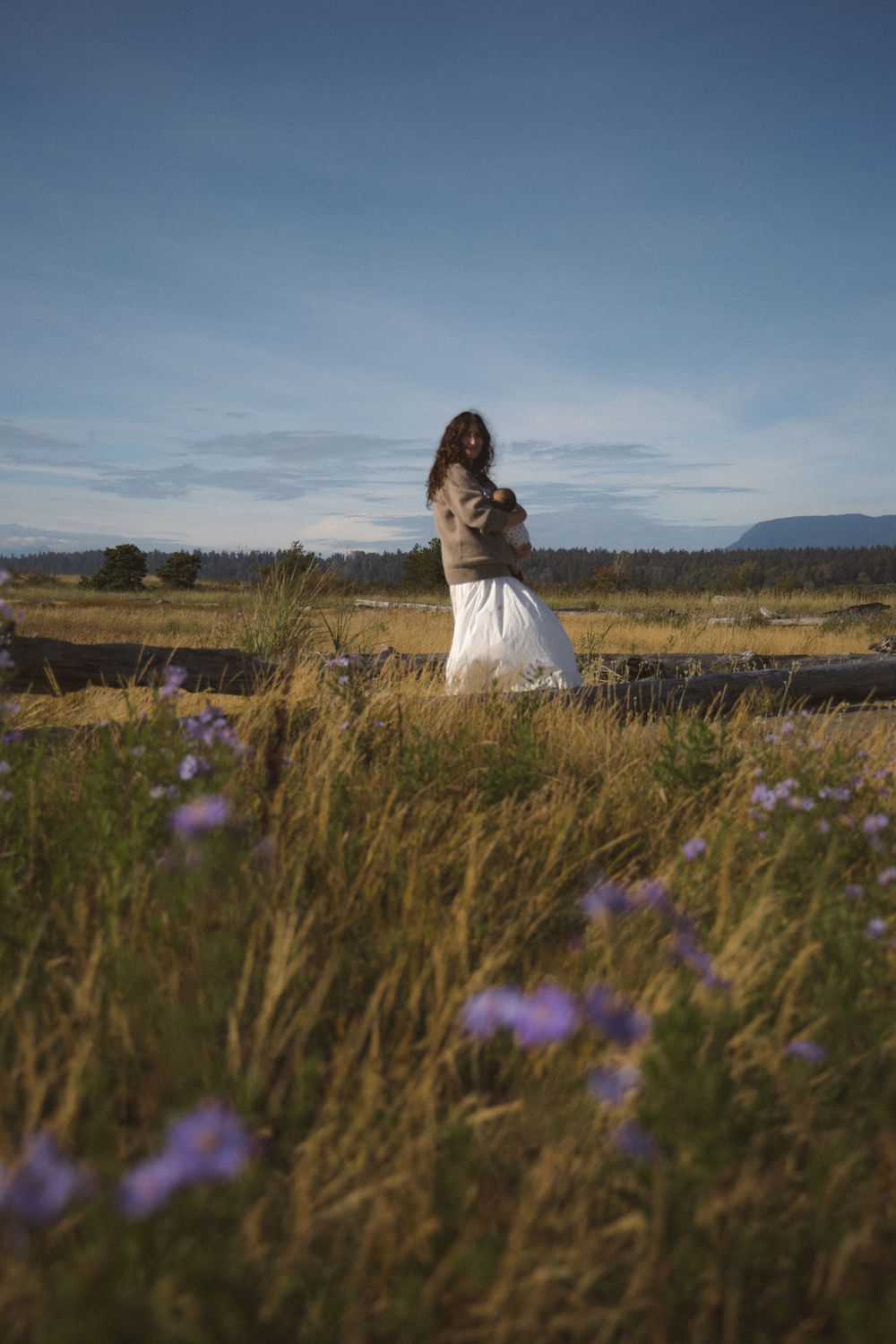 Mom holding her newborn baby in a field of purple flowers with blue skies above her during a newborn photoshoot in the outdoors in Vancouver