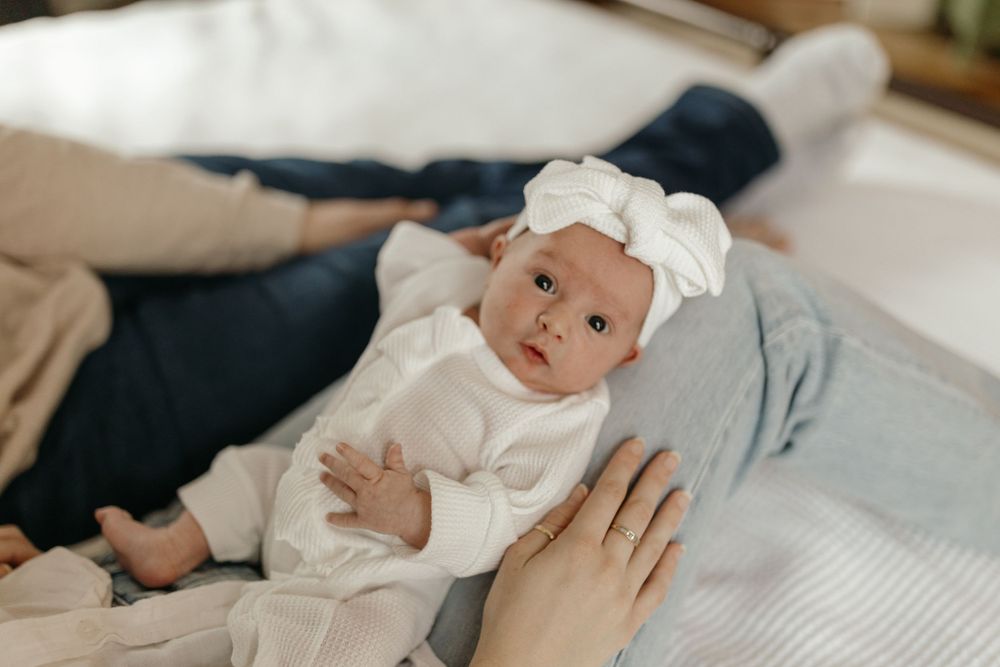 Baby wearing white outfit and bow lies on bed while being gently touched by adult hand. Virginia/ North Carolina Newborn In-Home Session
