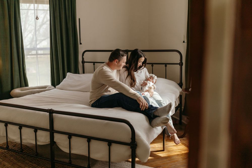 Cozy bedroom scene with couple sitting on metal frame bed by window with green curtains. Virginia/ North Carolina Newborn In-Home Session
