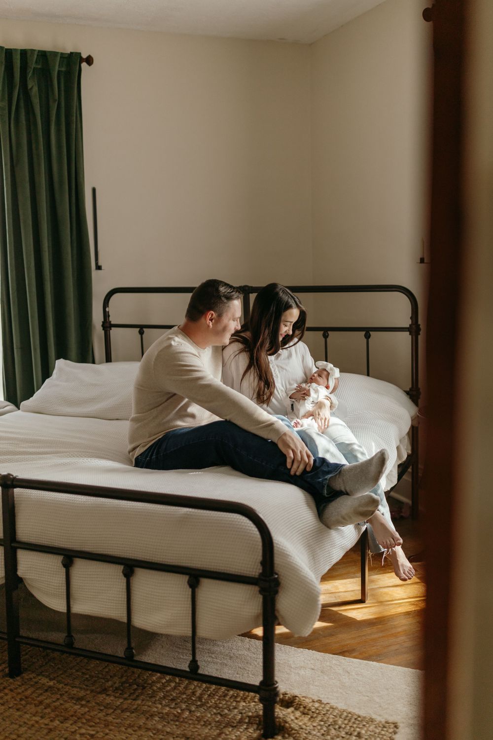 Family shares intimate moment together on metal frame bed near window with green curtains. Virginia/ North Carolina Newborn In-Home Session