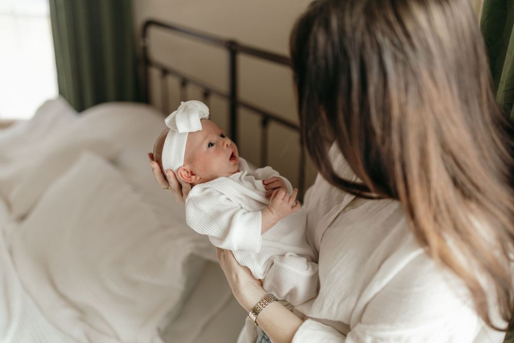Newborn baby wearing white outfit and bow being held on white bedding. Virginia/ North Carolina Newborn In-Home Session