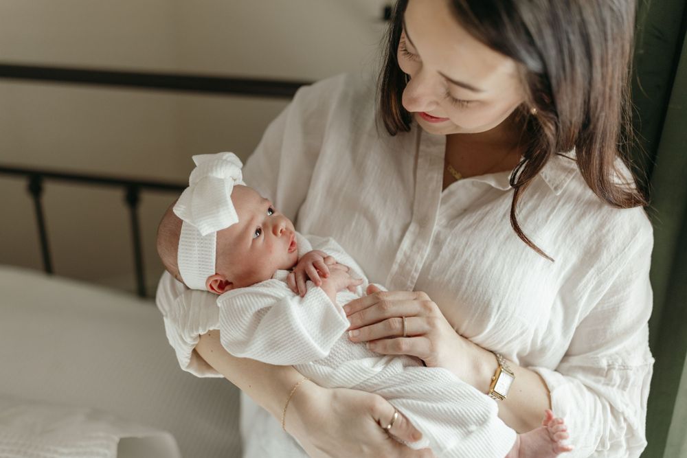 Gentle moment with newborn in white outfit being held on bed. Virginia/ North Carolina Newborn In-Home Session