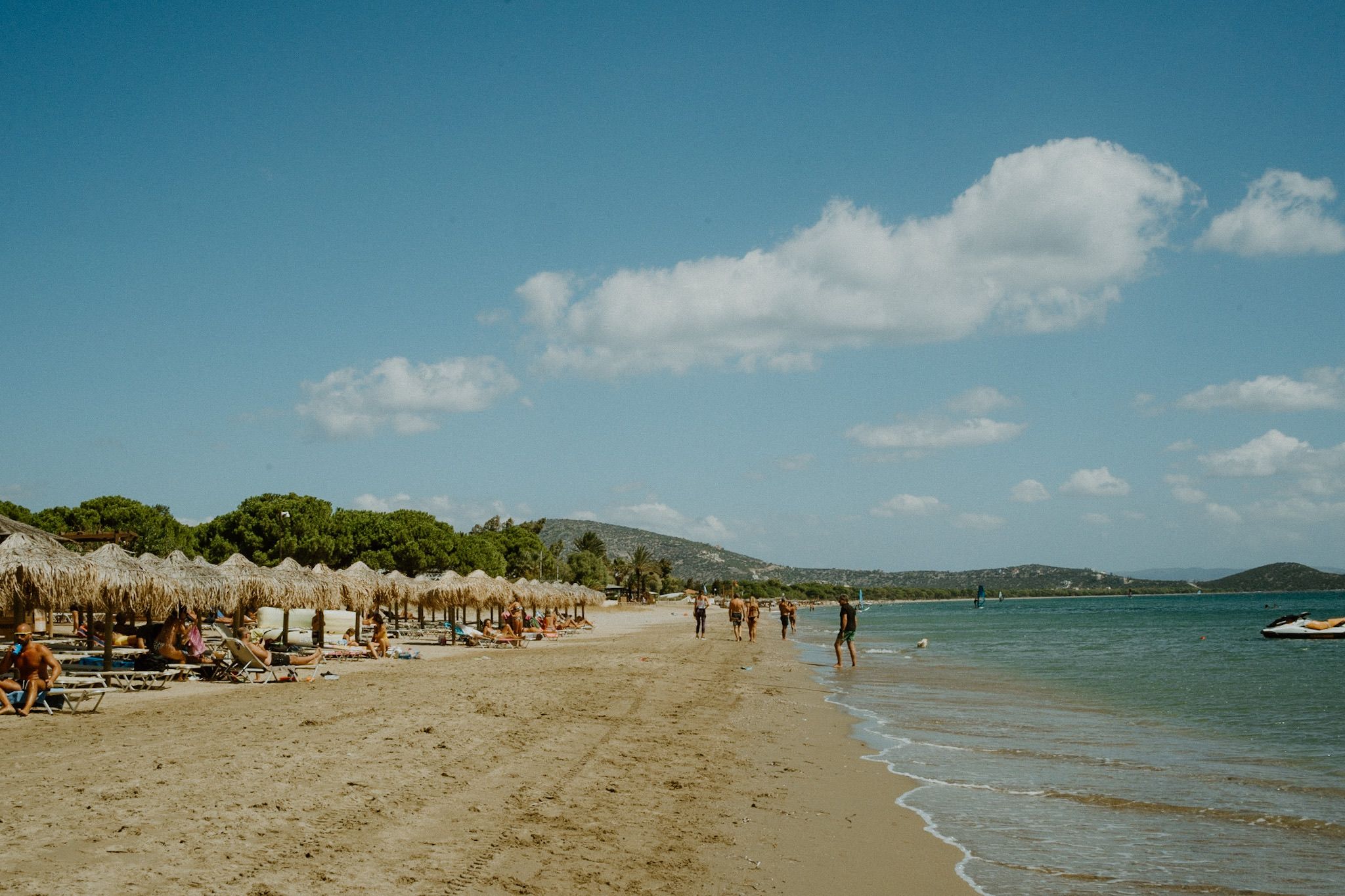 Tropical beach with thatched umbrellas and sunbathers on sandy shore under blue sky and white clouds.