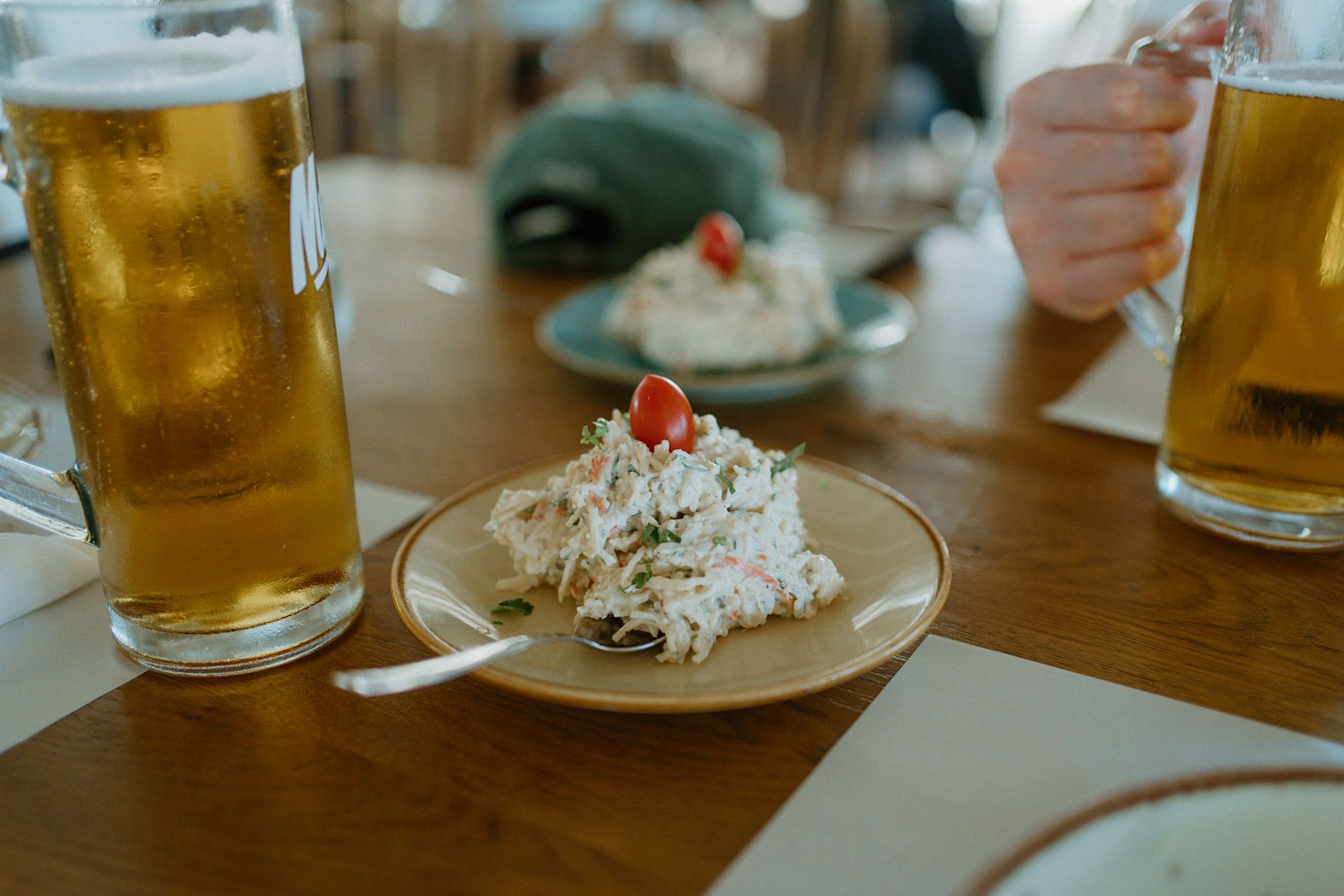 Two glasses of cold beer with a small plate of crab salad garnished with a cherry tomato on a wooden restaurant table.