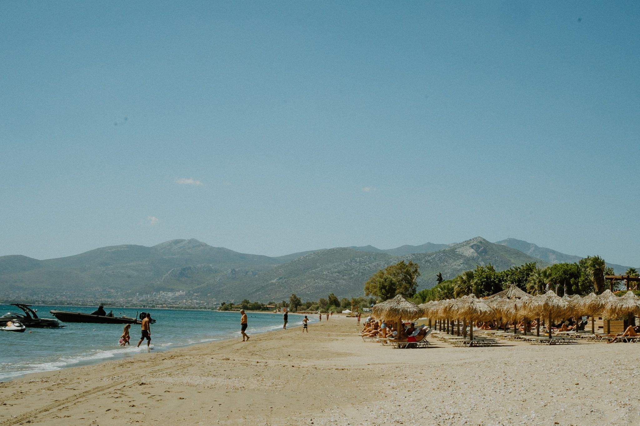 Scenic Mediterranean beach with thatched umbrellas, mountains in background and beachgoers enjoying sunny day.