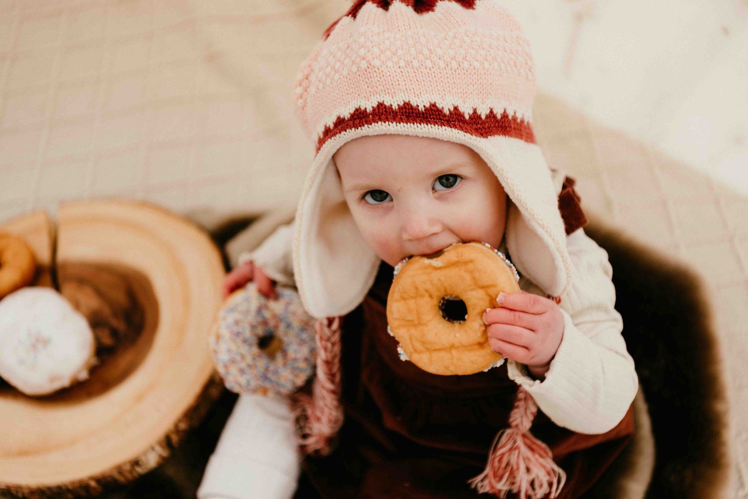 A child in a winter hat takes a bite of a glazed donut while sitting near a wooden plate of pastries.