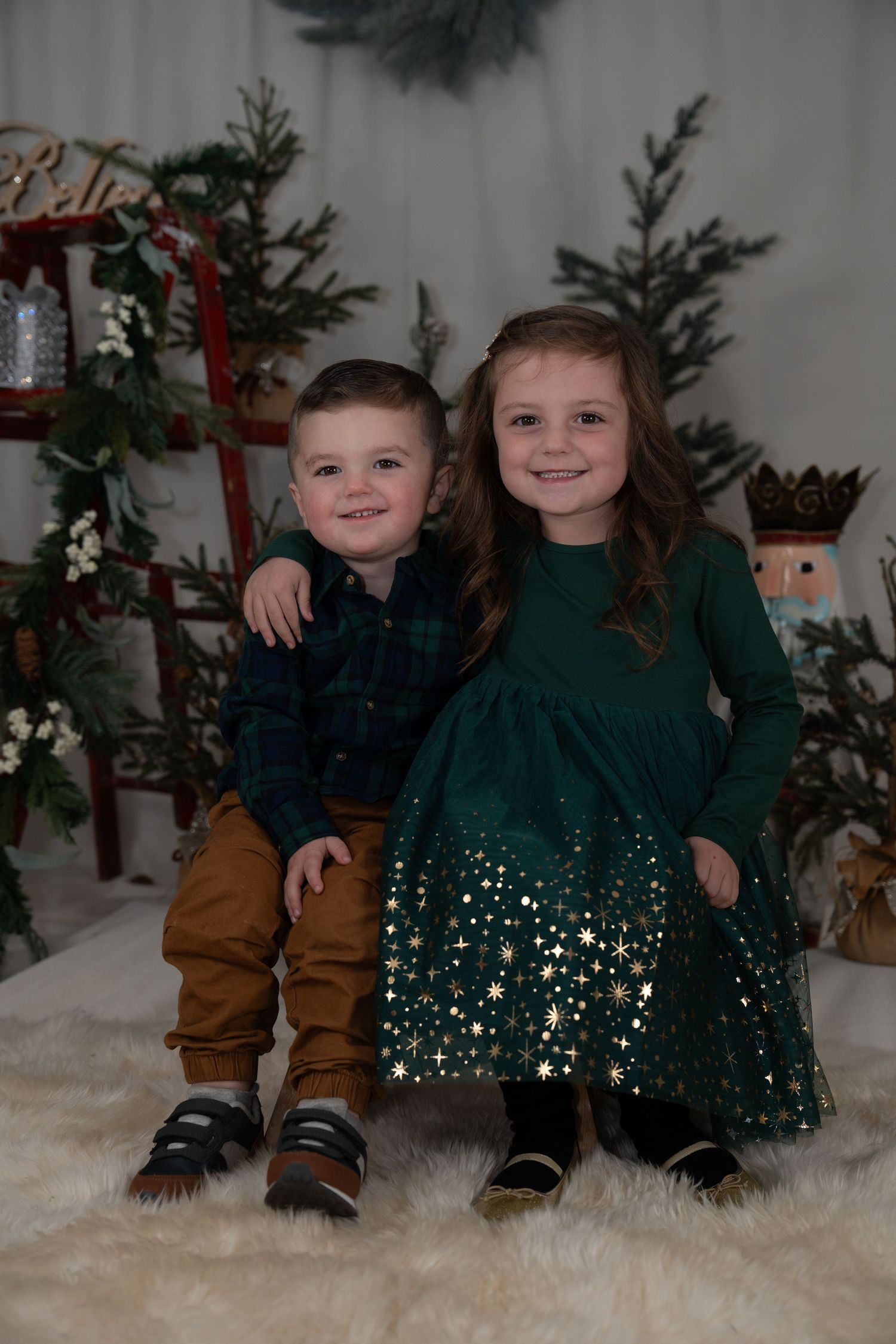 Children in matching green holiday outfits sitting on white fluffy rug near Christmas trees.