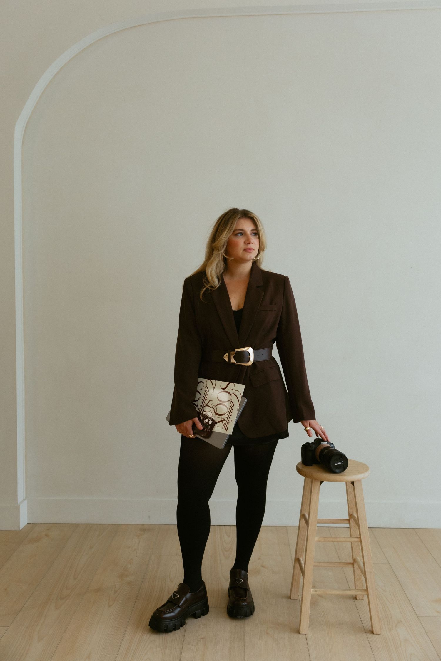 Woman in gray blazer and black outfit sits cross-legged working on laptop with camera nearby on white background.