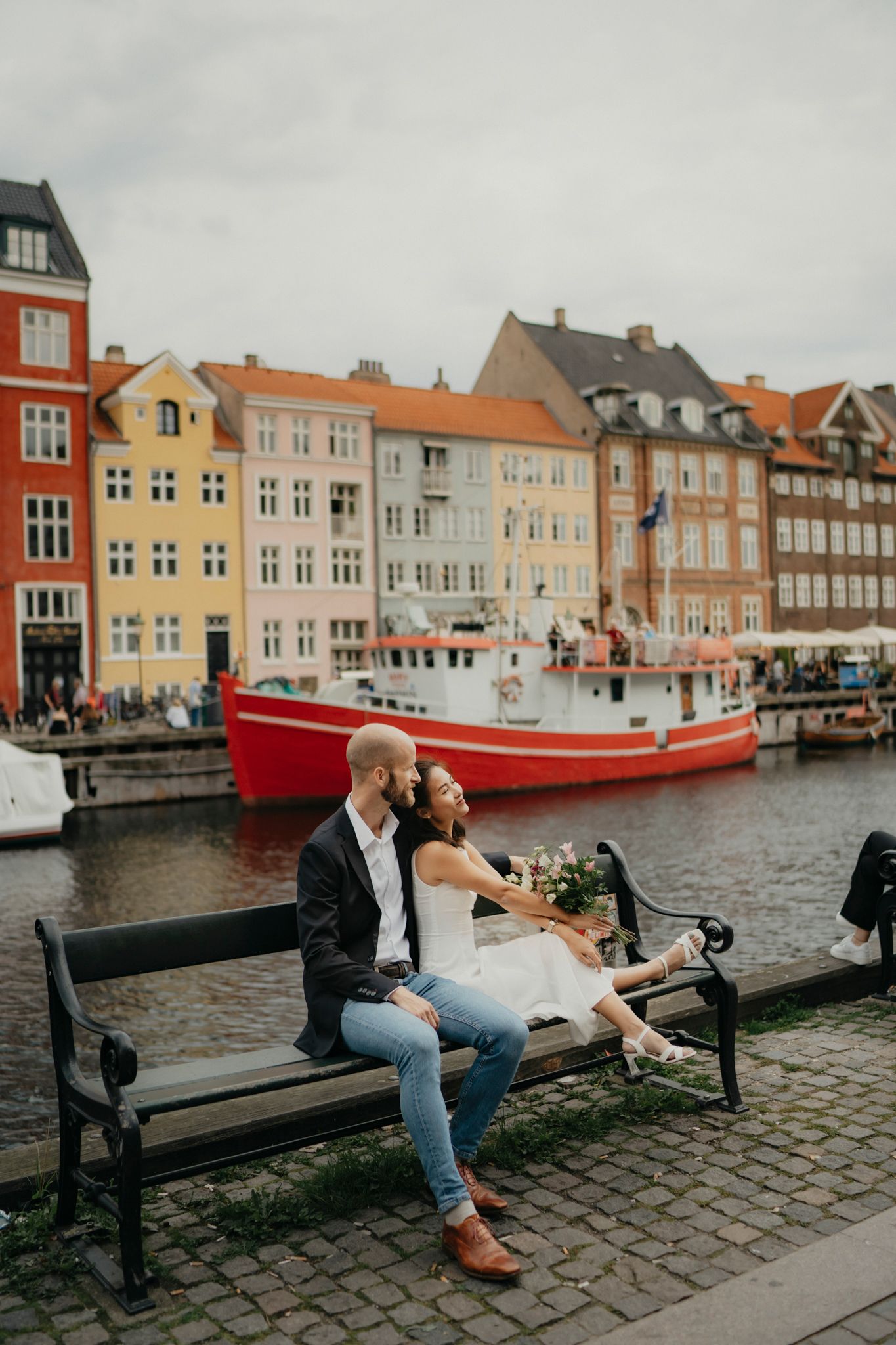 Couple sitting on bench by harbor with colorful Danish buildings and red boat in background.