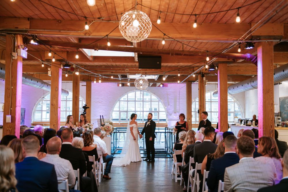 Wedding ceremony in progress in an industrial-style venue with wooden beams and string lights.