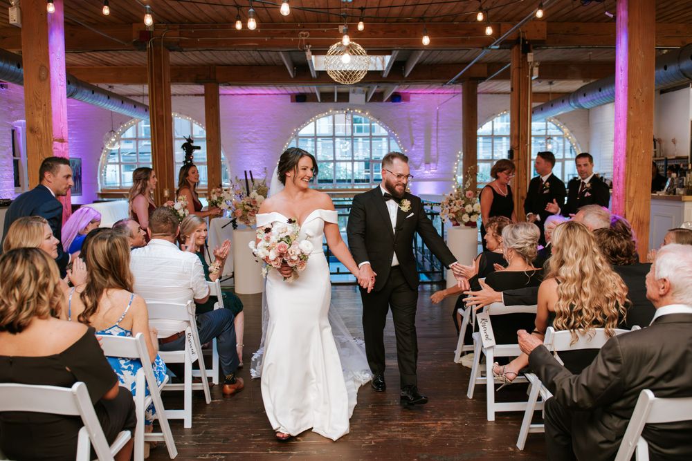 Wedding ceremony exit in an industrial chic venue with purple accent lighting and white chairs.