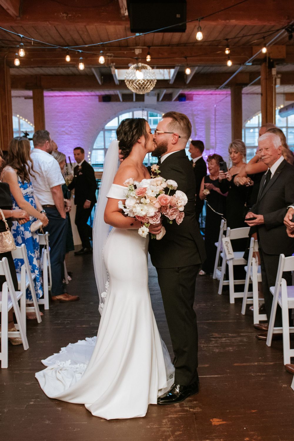 Newlyweds share their first kiss in an elegant venue with purple uplighting and exposed wooden beams.