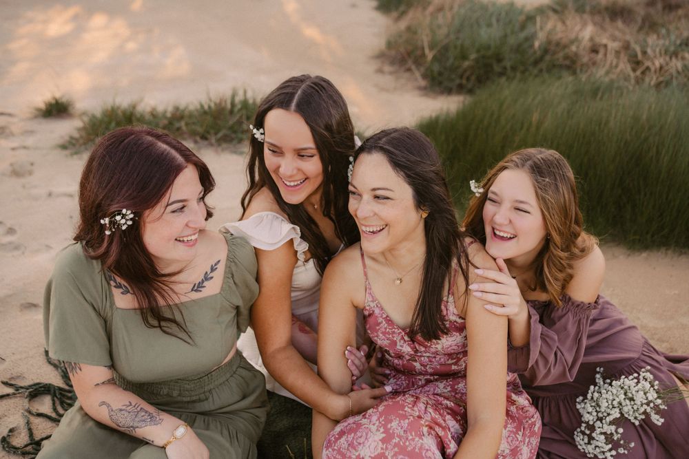 Four friends share joyful laughter while sitting together outdoors at sunset with dried flowers and natural scenery.
