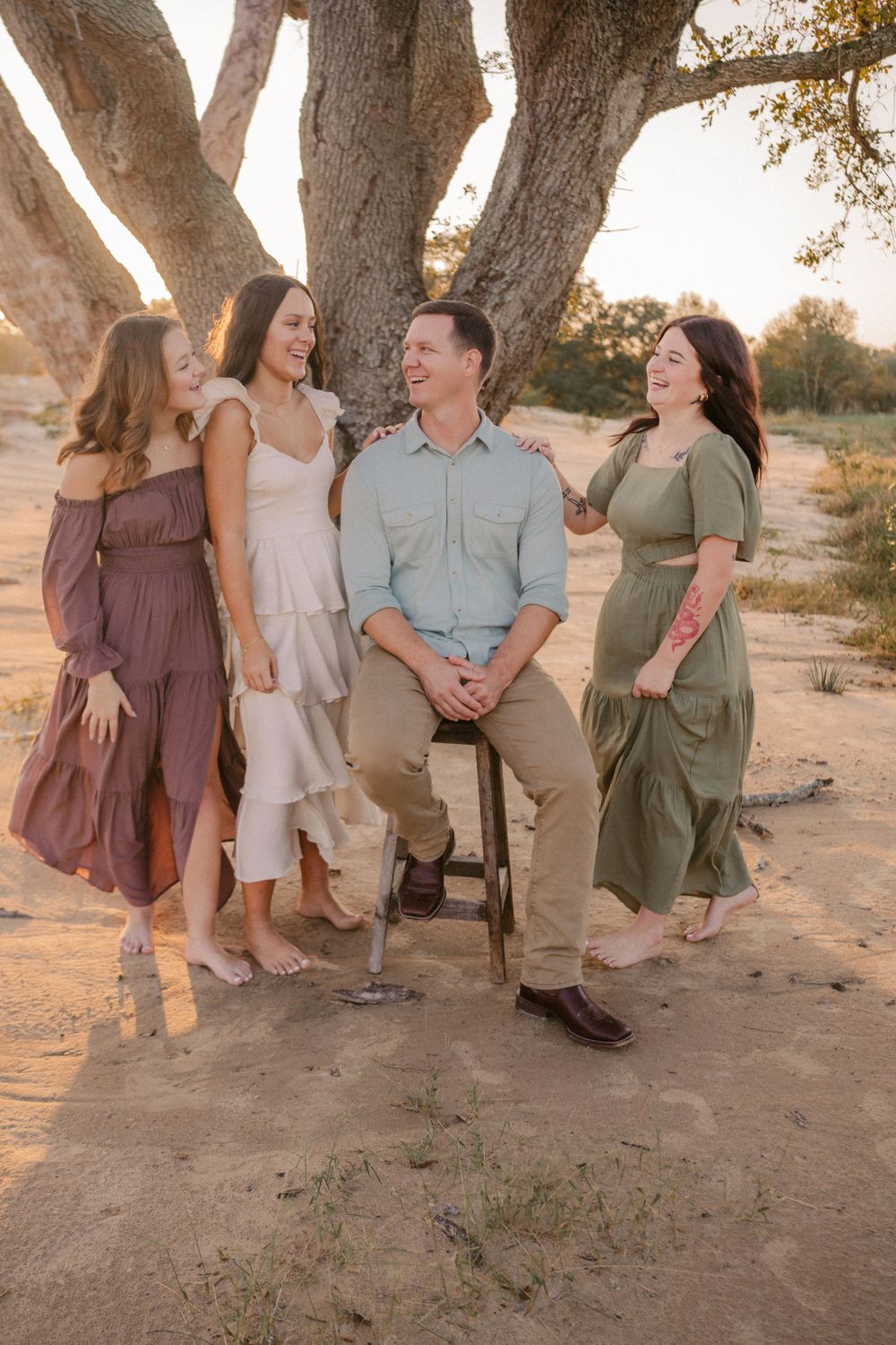 A group shares joyful laughter while posing together under large trees at sunset in casual dressy outfits.