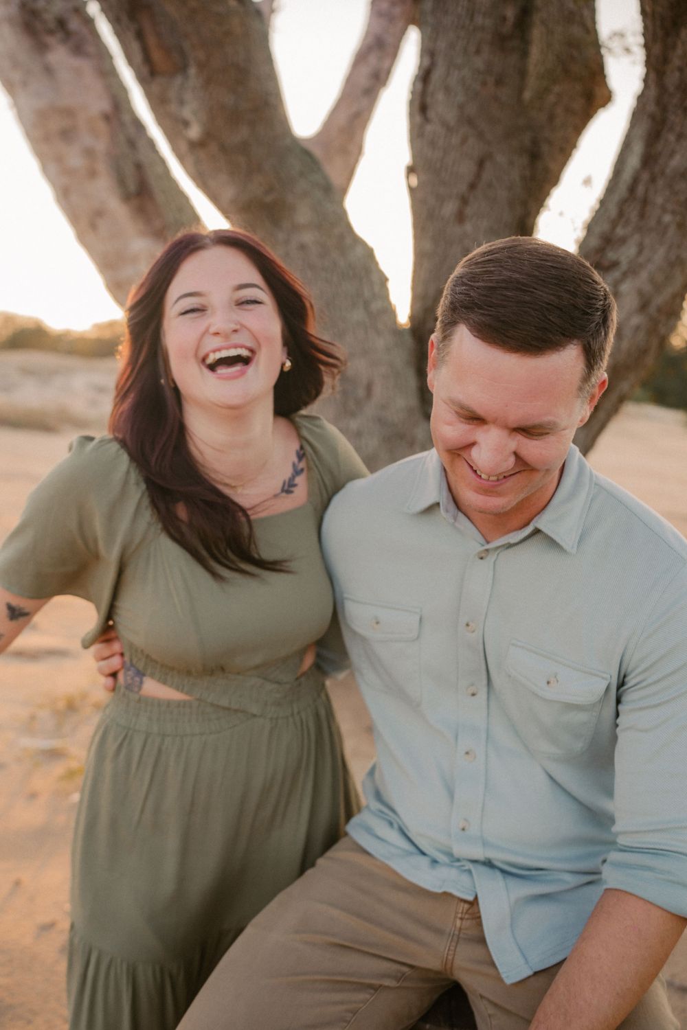 Happy couple laughing together outdoors on a sunny day with a rustic tree backdrop.