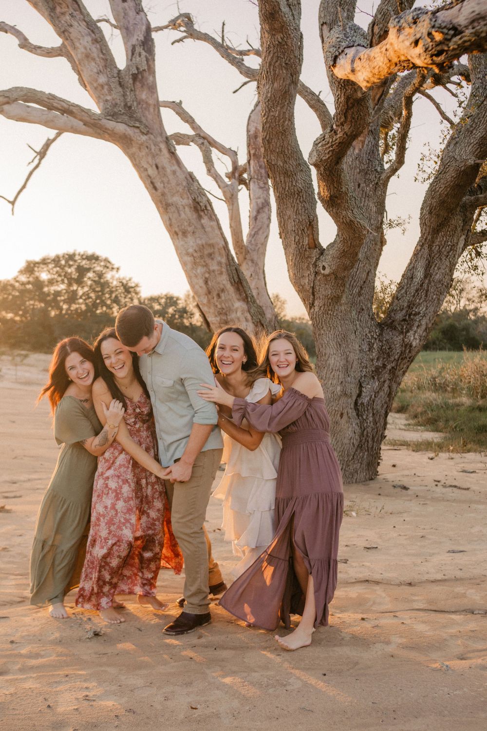 A group laughs together under a large tree at sunset while wearing flowing dresses in earth tones.