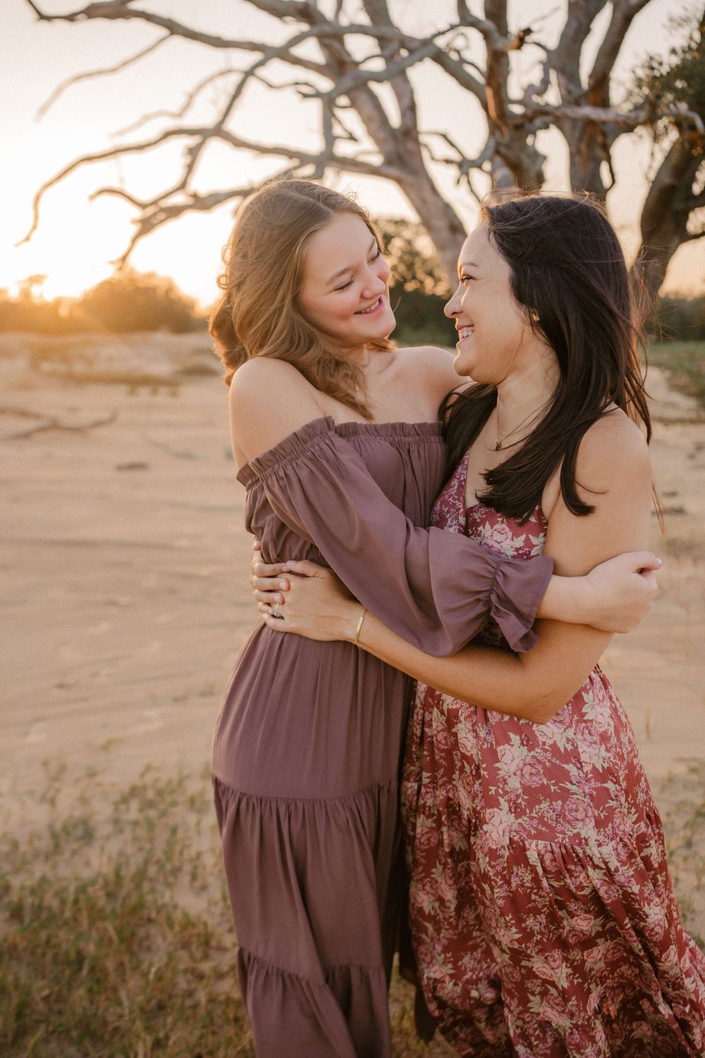 Two friends share a joyful embrace at sunset in a desert landscape with bare tree branches overhead.