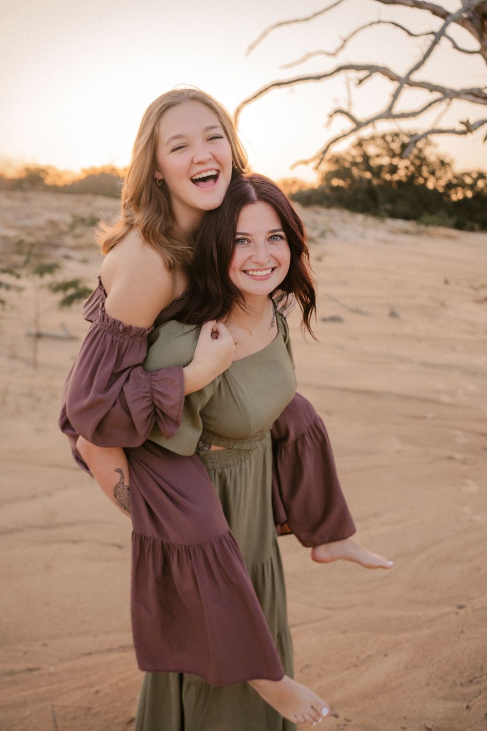 Two friends share joyful moment in desert landscape at sunset while wearing flowy dresses and embracing.