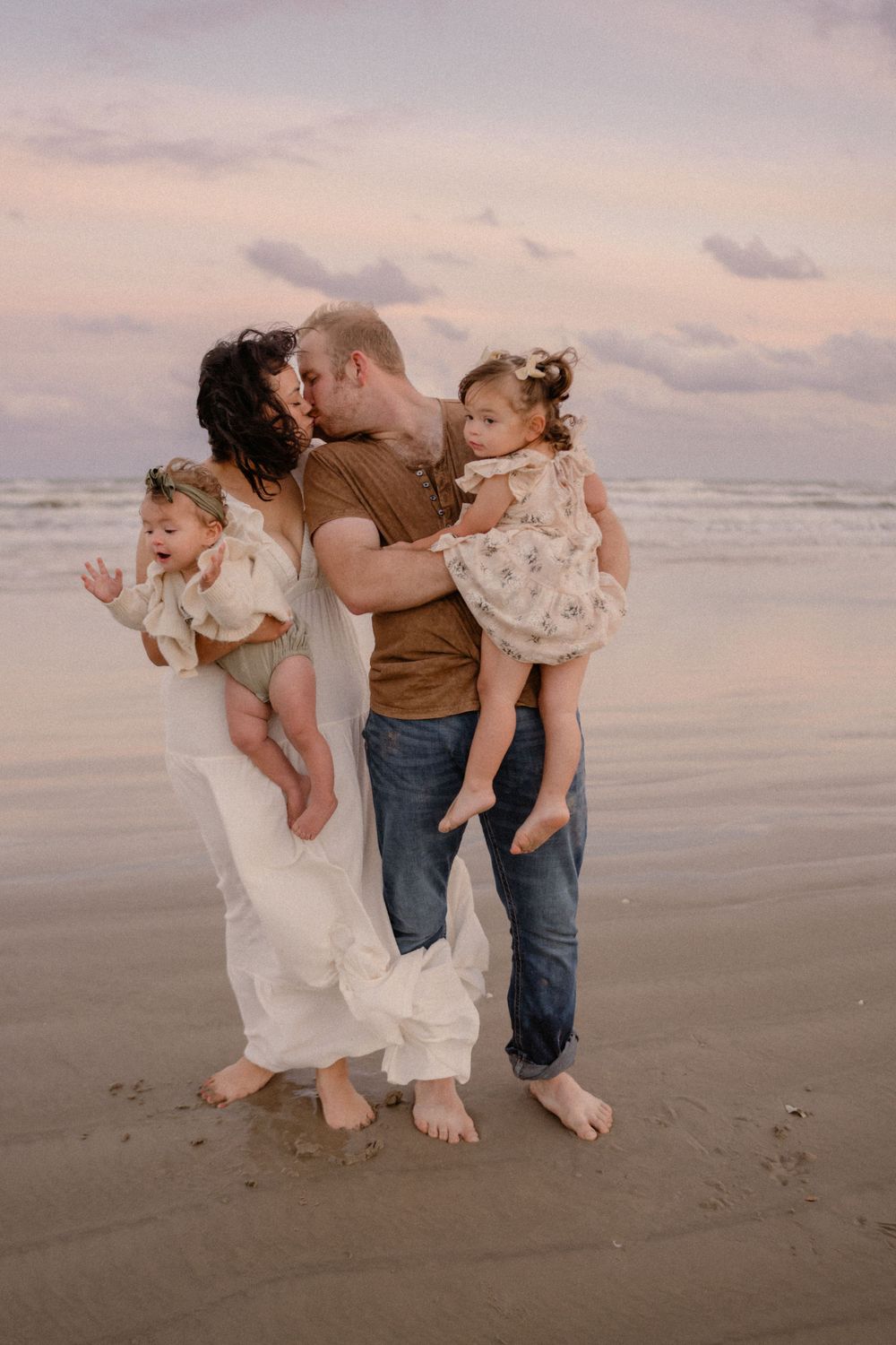 Family shares tender moment together on a sandy beach at sunset with gentle waves in the background.
