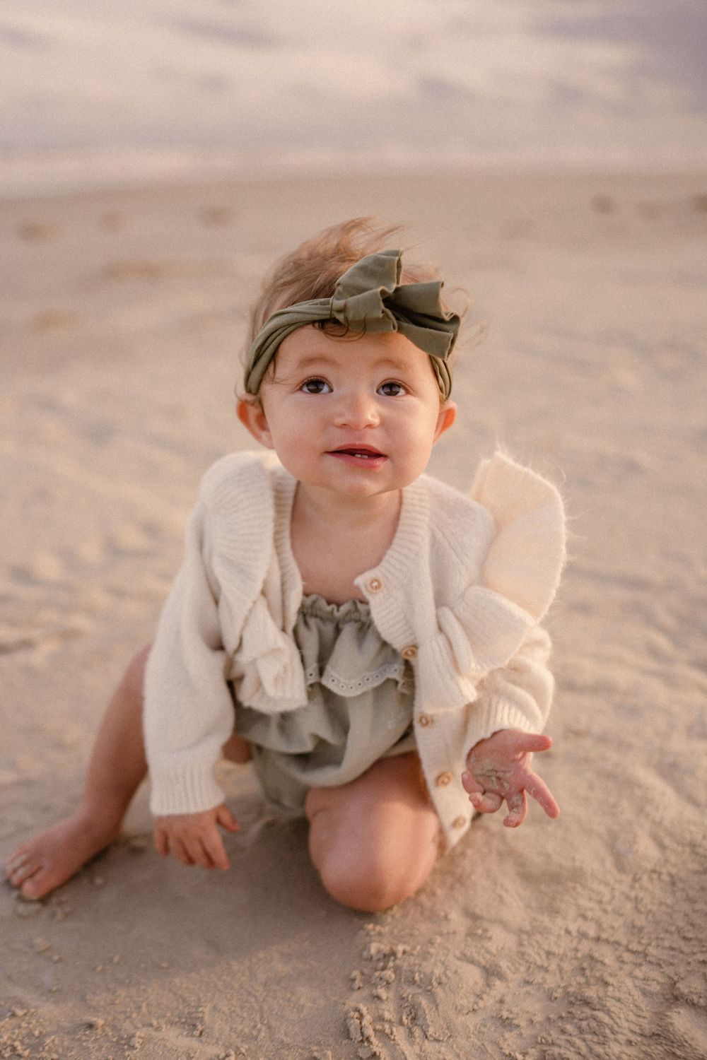 Small child wearing gray outfit and white cardigan kneels and plays on sandy beach at sunset.