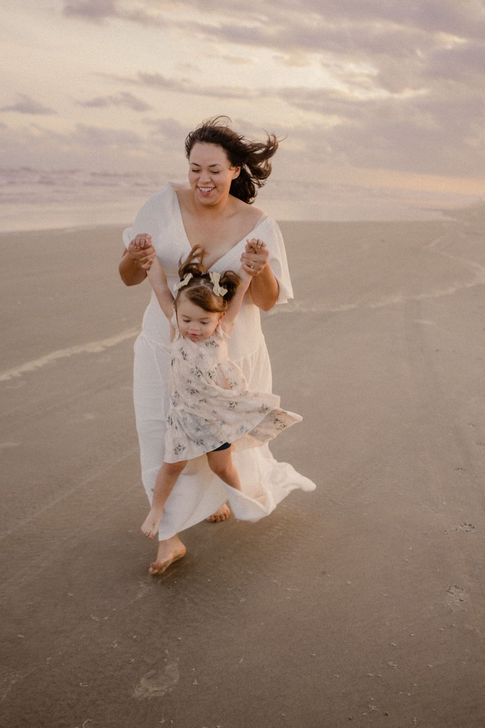 A joyful moment captured on a cloudy beach as a mother and child in white dresses run together on the damp sand.