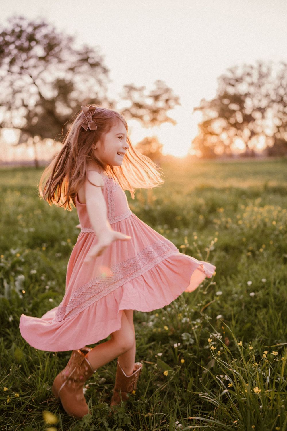 A pink-dressed figure twirls joyfully in a wildflower meadow at sunset with hair flowing in the golden light.