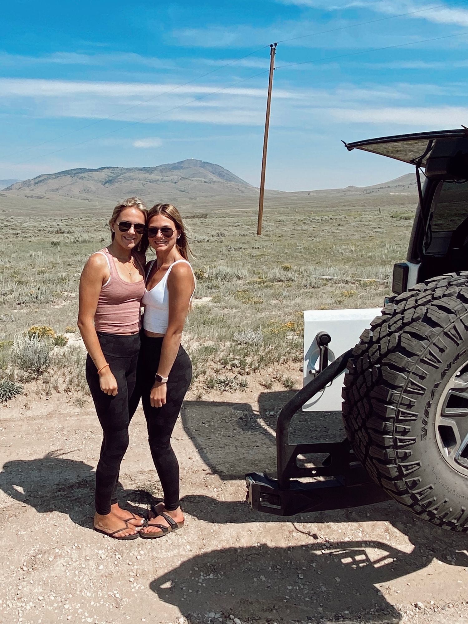 Two people pose beside a 4x4 vehicle in a vast desert landscape with mountains and utility poles in the distance.