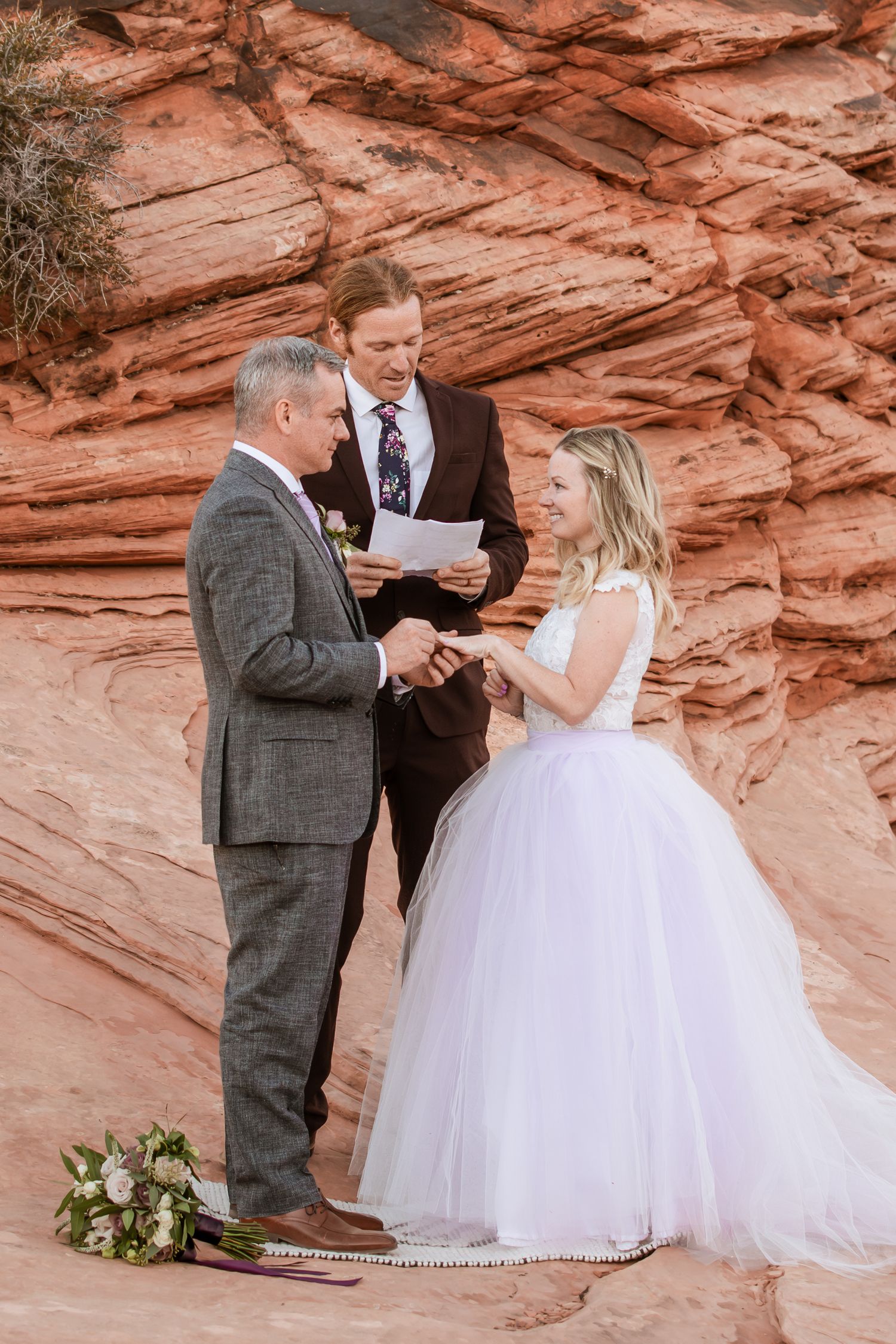 A wedding ceremony takes place against striking red sandstone cliffs with the couple and officiant exchanging vows.