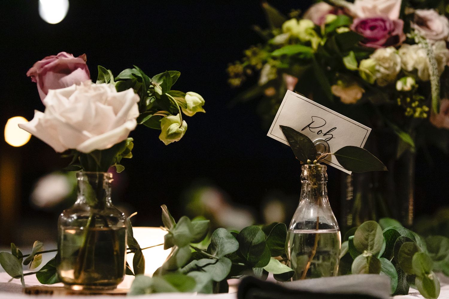 Elegant table setting with pink rose in glass bottle vase and greenery accents.