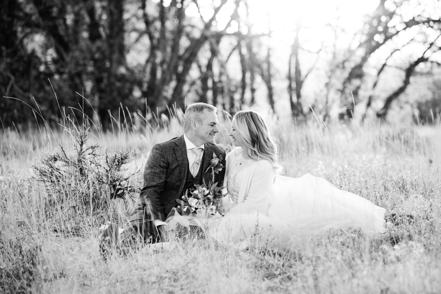 A romantic black and white portrait of newlyweds sitting together in a meadow.
