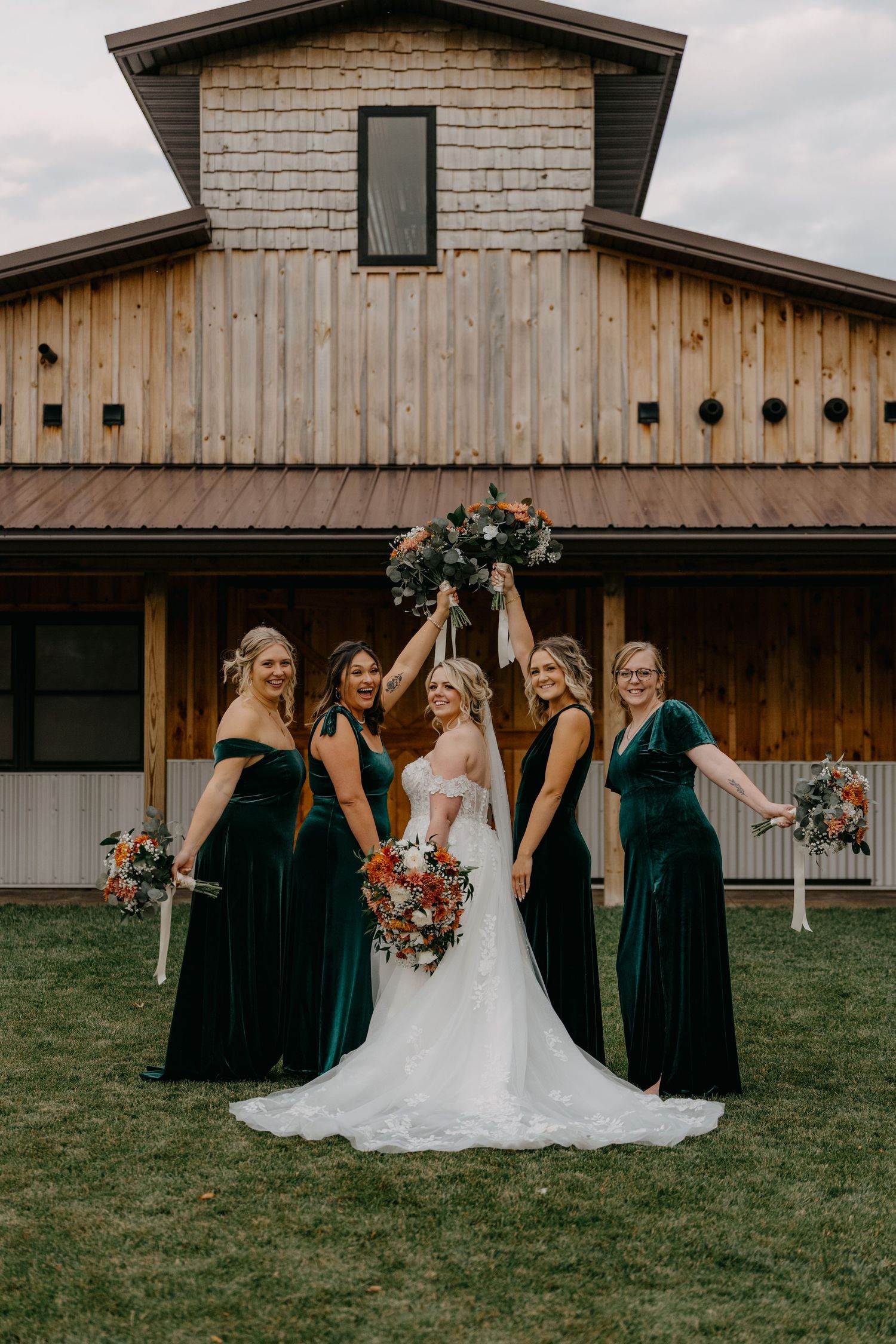 Wedding party in emerald velvet bridesmaid dresses poses together on lawn in front of rustic wooden barn venue.
