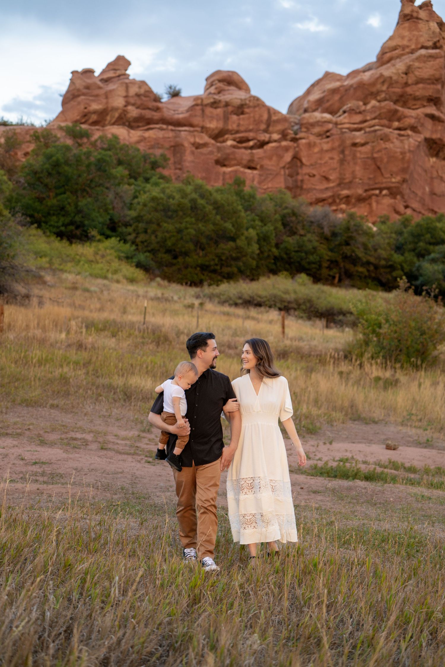 A couple stands together with their baby against red rock formations at sunset in a scenic outdoor setting.