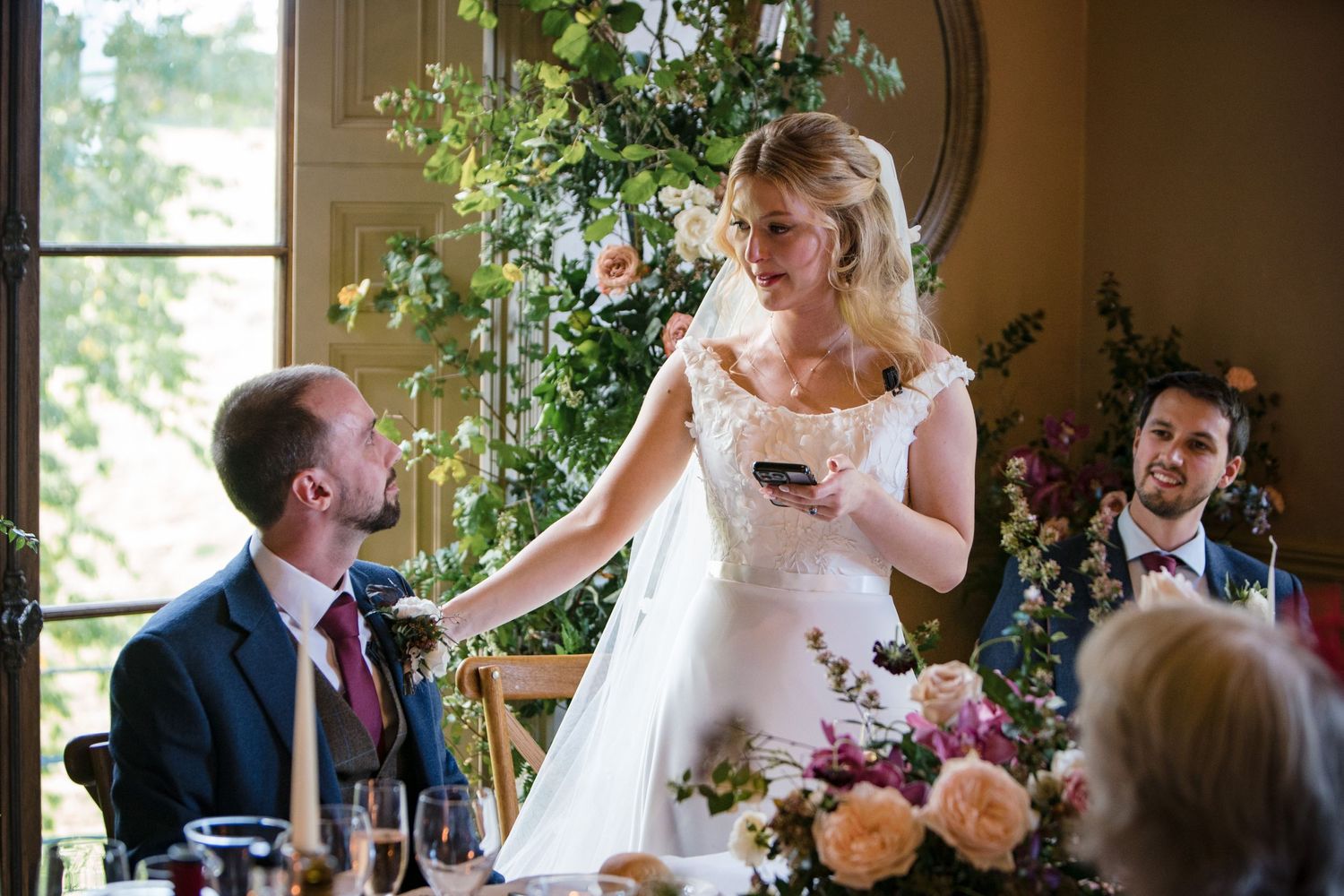 A bride in a wedding dress delivers a speech at Wilson Coastal Club, a reception table decorated with floral arrangements.