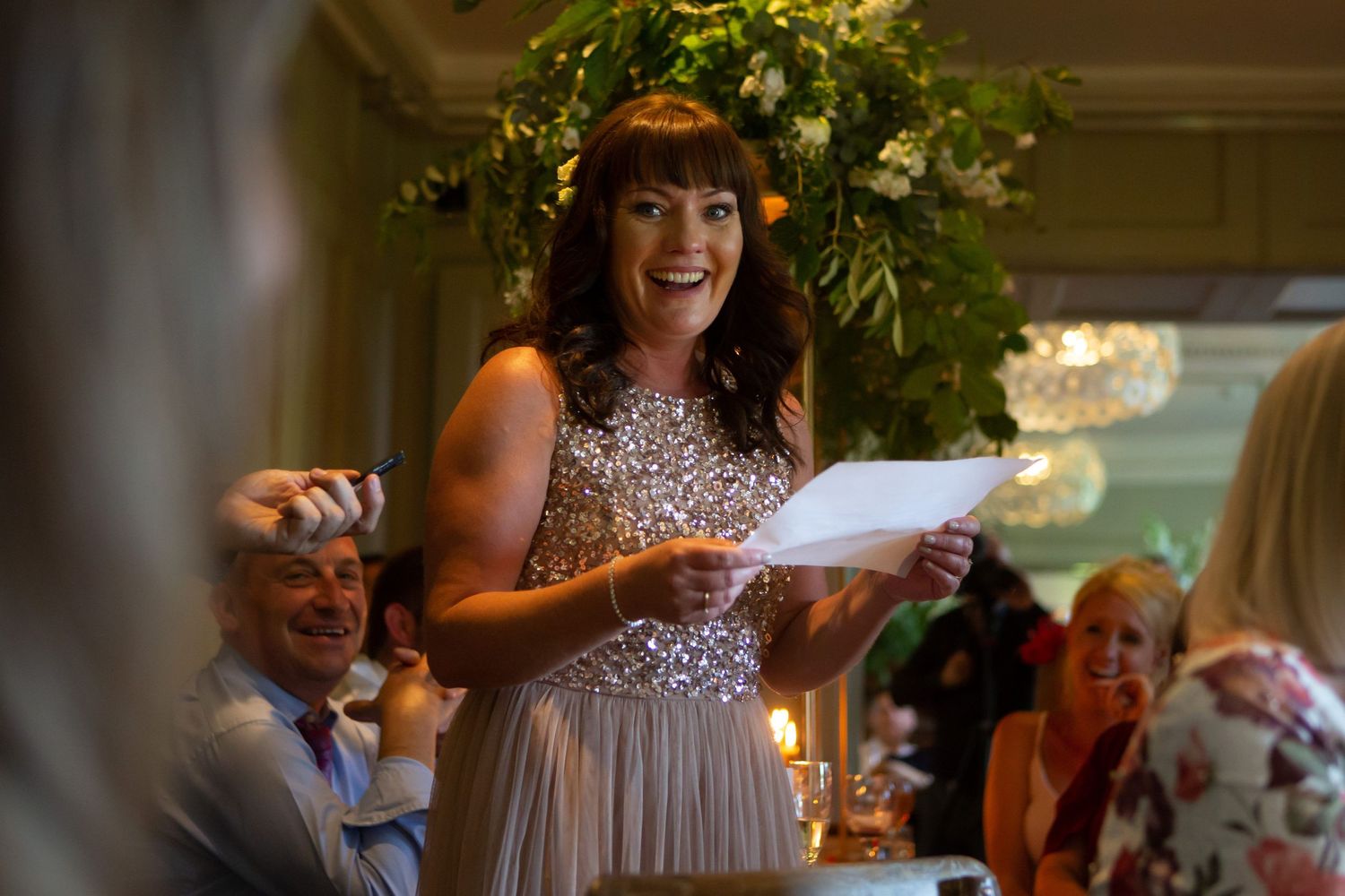 A bridesmaid in a sparkly dress gives a speech during a wedding reception at Wilson Coastal Club while guests look on and smile.