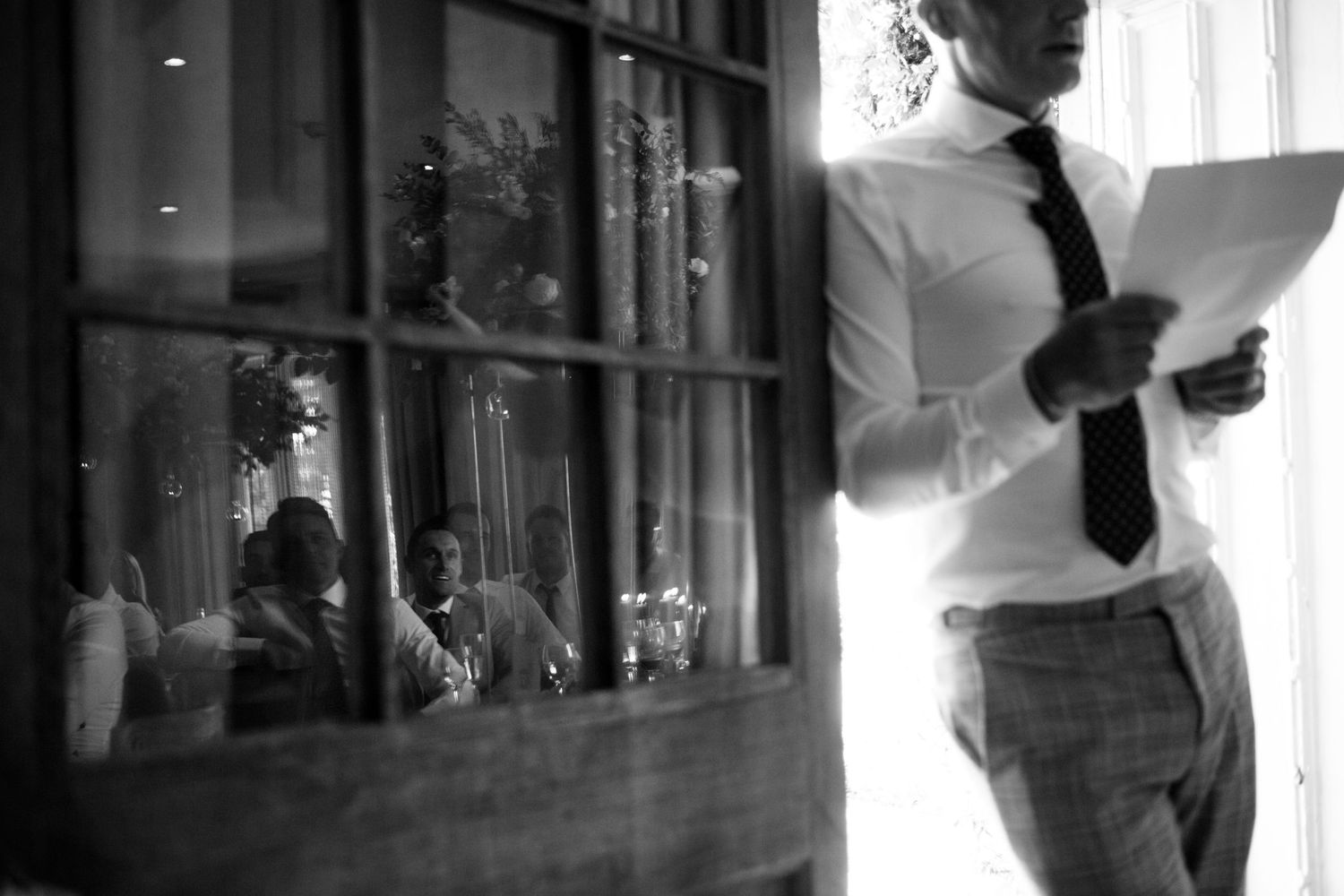 A black and white photograph of Bestman in formal wear reading speech next to a window at Wilson Coastal Club wedding venue in Nova Scotia.