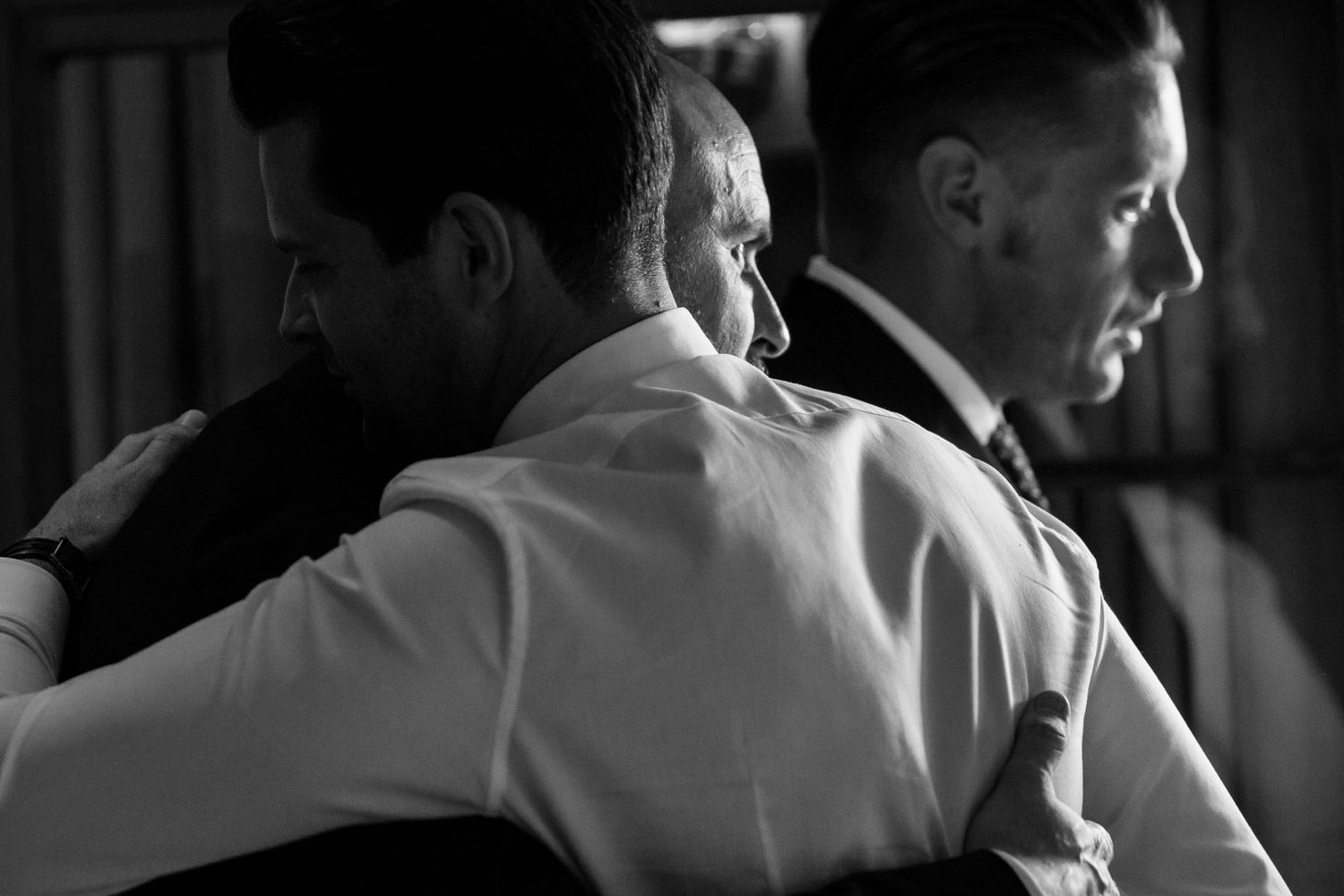 A black and white candid photo showing two men in formal attire hugging in Wilson Coastal Club wedding venue.