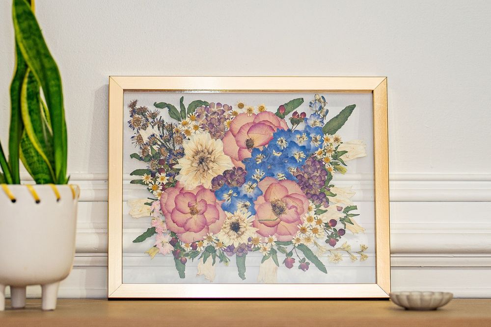 Framed floral bouquet artwork with pink, blue, and yellow flowers in a gold frame on a wooden shelf.