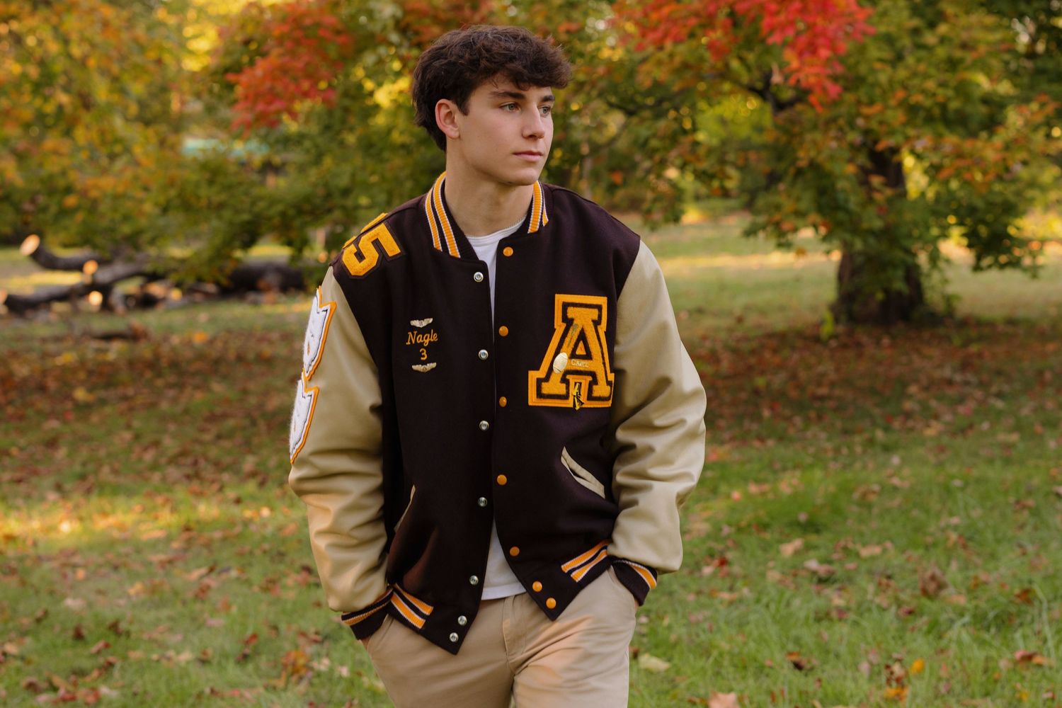 A classic varsity letterman jacket in brown and cream colors photographed against autumn foliage in a park setting.