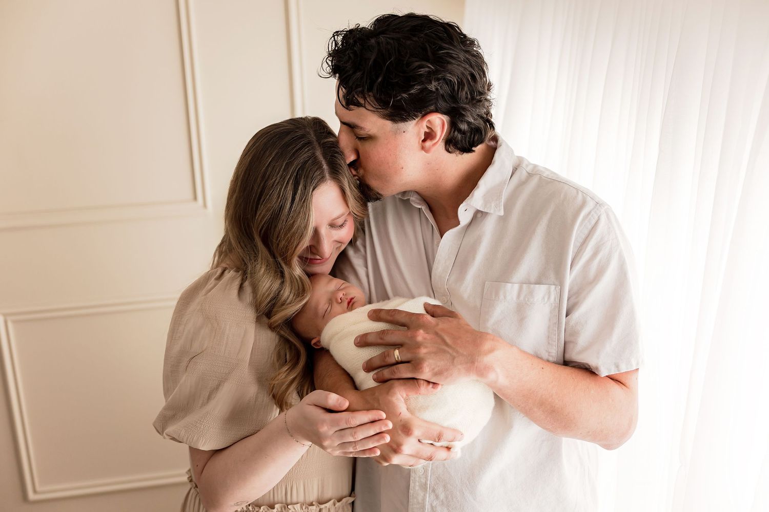 A tender family portrait shows parents in neutral tones embracing their newborn baby while an older sibling leans in close.