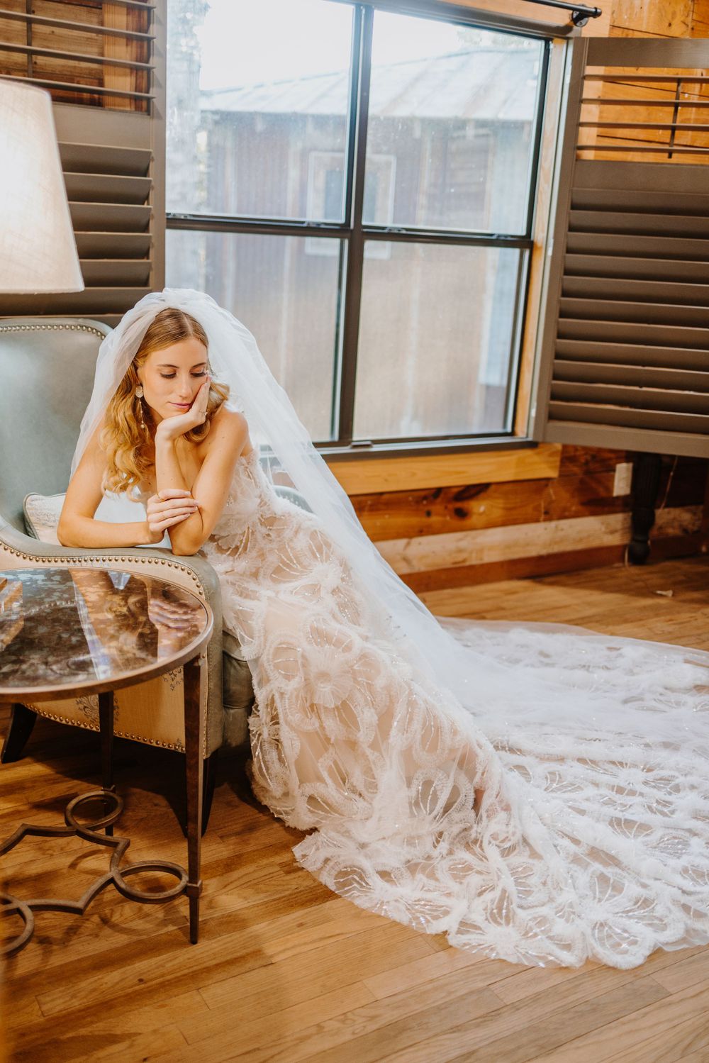 Bride in flowing lace gown poses elegantly on wooden floor near window.
