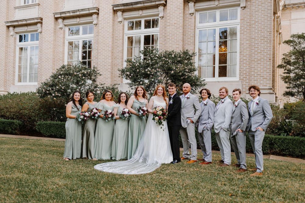 Wedding party poses outside a grand brick building in formal attire with bridesmaids in sage green dresses.