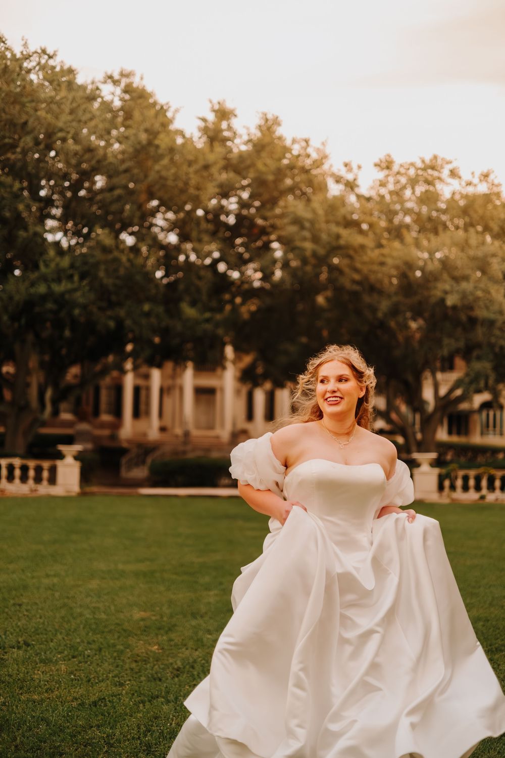 A bride in a strapless white gown twirls joyfully on a sunlit lawn in front of a grand estate.